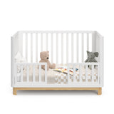 Front view of a white crib with a natural wood color base, featuring guard rails in toddler bed conversion